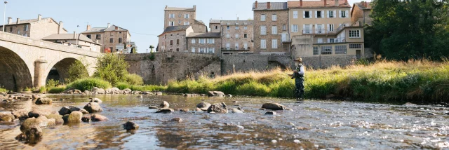 Au premier plan, on voit la rivière du Lignon qui coule avec un pêcheur à la mouche. Au second plan, un pont enjambe la rivière et les maisons alignées du village de Tence apparaissent serrées les unes contre les autres