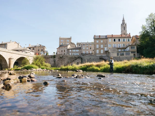 Au premier plan, on voit la rivière du Lignon qui coule avec un pêcheur à la mouche. Au second plan, un pont enjambe la rivière et les maisons alignées du village de Tence apparaissent serrées les unes contre les autres